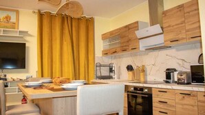 Une cuisine moderne avec des meubles en bois, un four encastré et un coin repas comprenant une chaise blanche et une table dressée avec des assiettes et un panier.