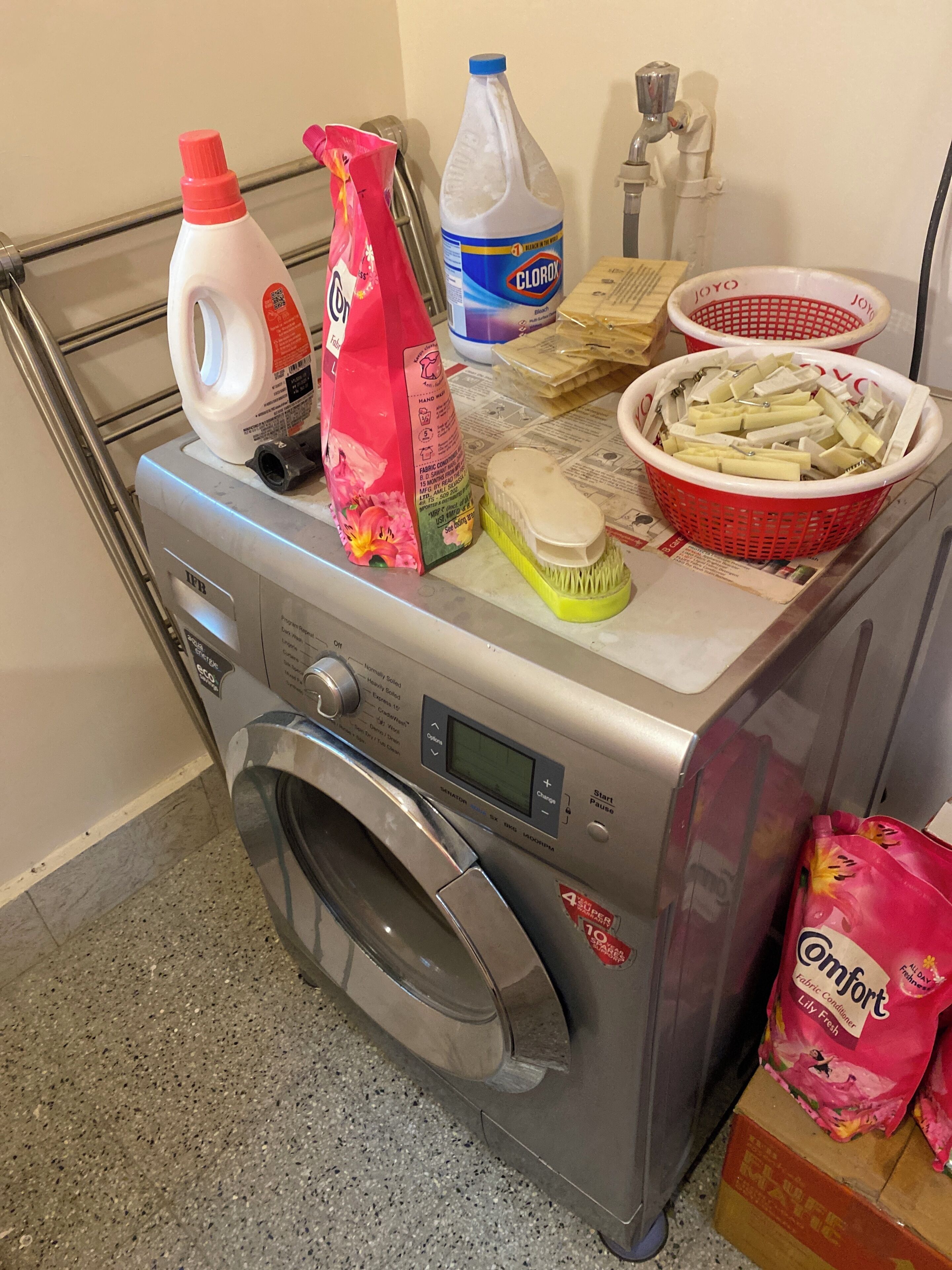 A front-loading washing machine with various laundry supplies on top, including detergent bottles, fabric softener, and a basket of clothes.
