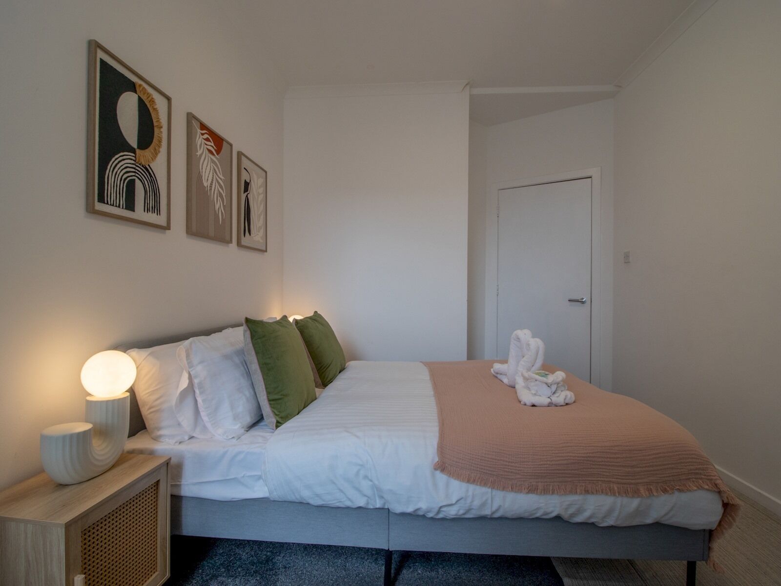 A neatly made bed with white and green pillows, a beige blanket, and a white towel on the footboard. There are framed artworks on the wall and a bedside lamp on a wooden nightstand.