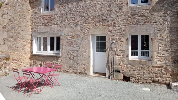 Un bâtiment en pierre avec une terrasse en gravier, une table et des chaises rouges, et une porte.