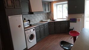 Une cuisine avec des meubles de rangement de couleur sombre, un lave-linge, un four à micro-ondes et une fenêtre avec des stores.