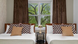 Une chambre d’hôtel avec deux lits, des coussins à carreaux, une petite table avec une lampe et une fenêtre donnant sur des arbres.