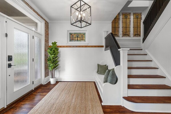 Un pasillo con piso de madera, un banco blanco con cojines, una planta en maceta, una lámpara de techo con diseño geométrico y una escalera con barandilla negra.