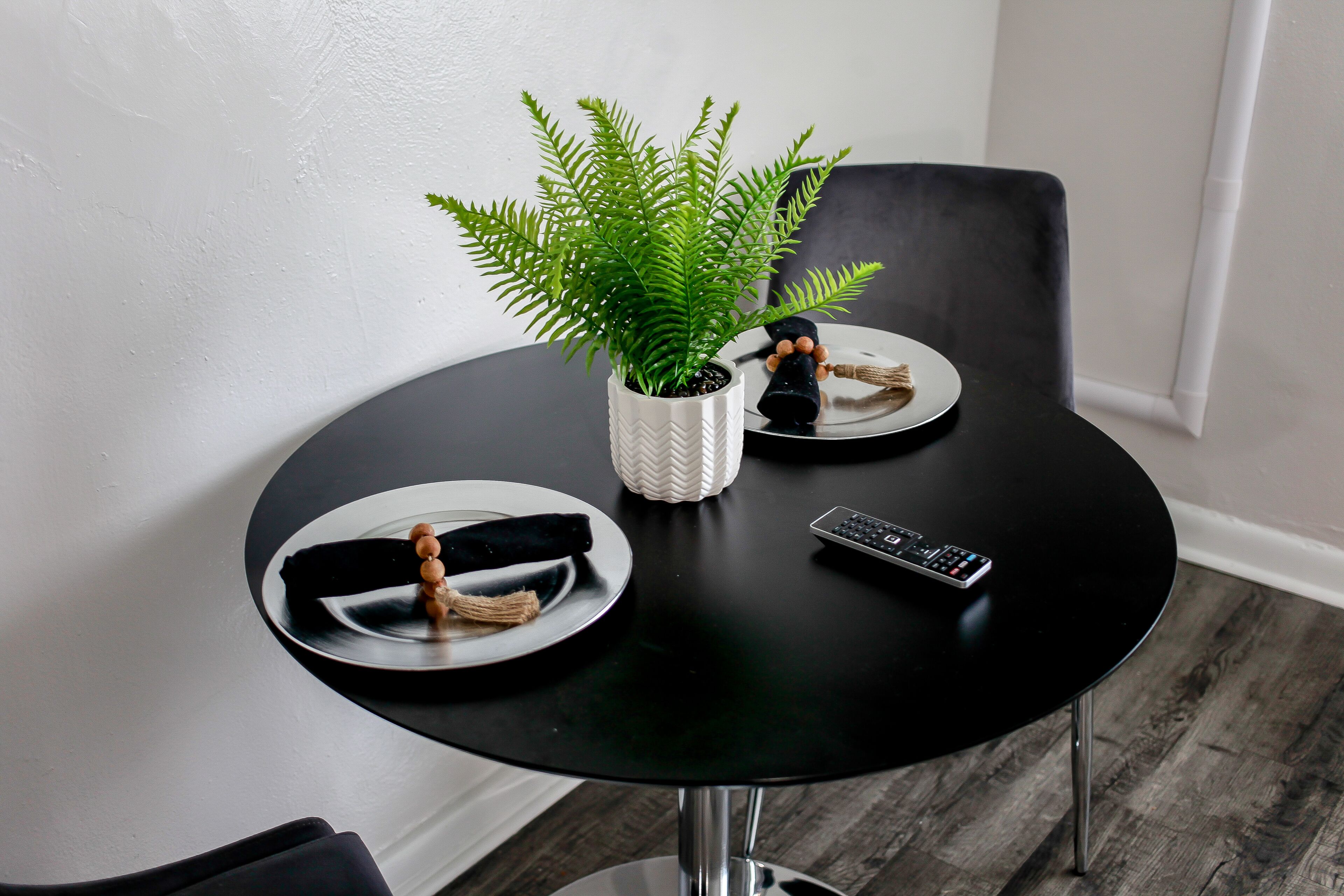A small round table with a potted fern, a remote control, and a plate with a black cloth and wooden beads.