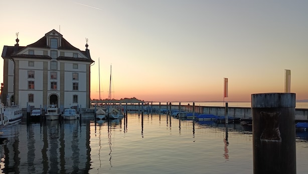 Un bâtiment situé au bord de l’eau, doté d’un style architectural classique, avec une étendue d’eau calme qui reflète sa façade, et un ciel dégagé au coucher du soleil.