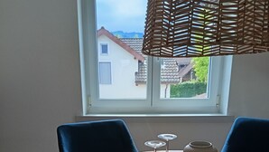 Une table ronde en bois, agrémentée de deux verres à vin et d’un vase, une lampe en osier et deux chaises bleues, dans une pièce dotée d’une fenêtre donnant sur une maison.