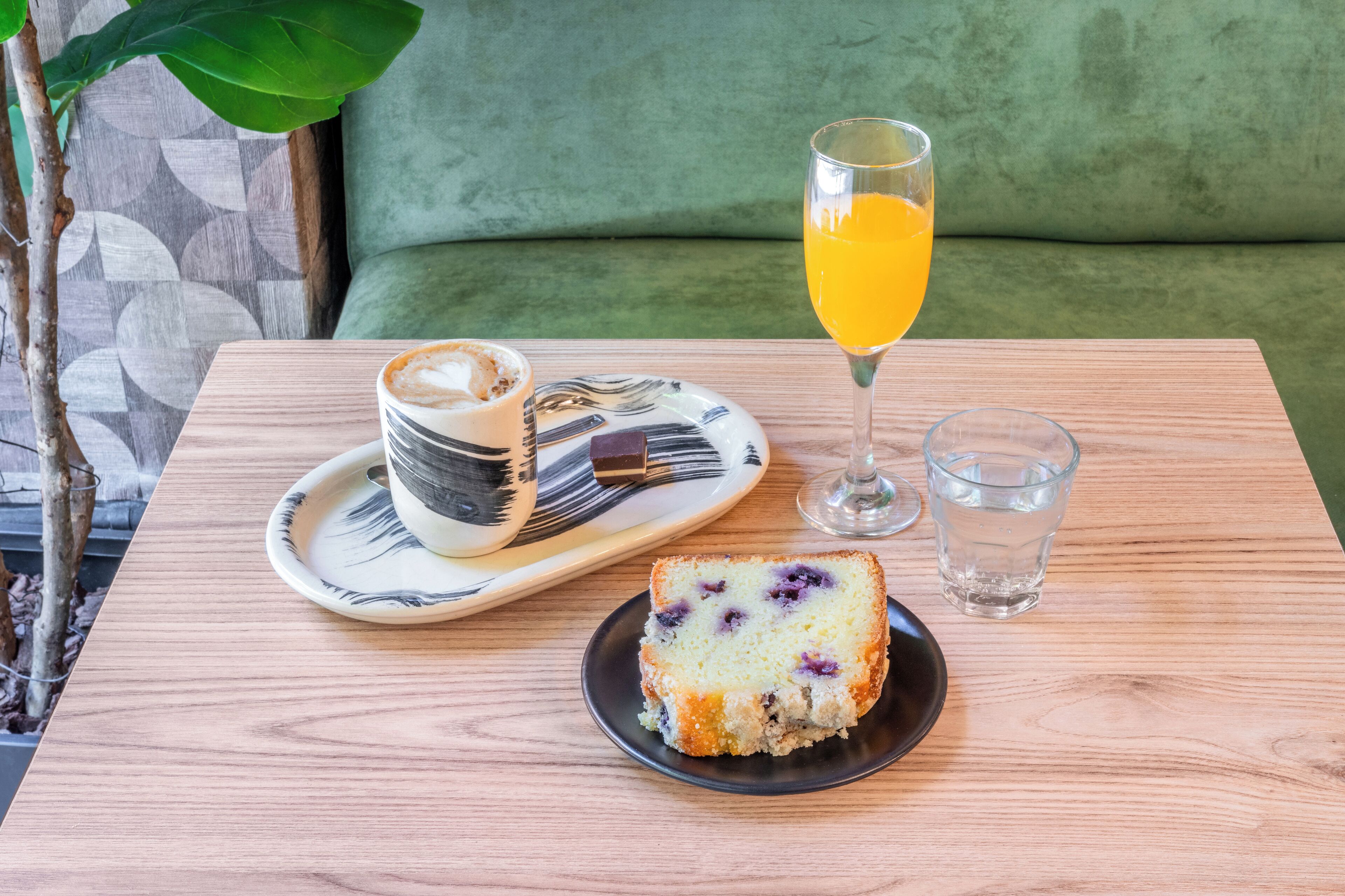 Een houten tafel met een plakje taart, een glas sinaasappelsap en een kop koffie op een bord.