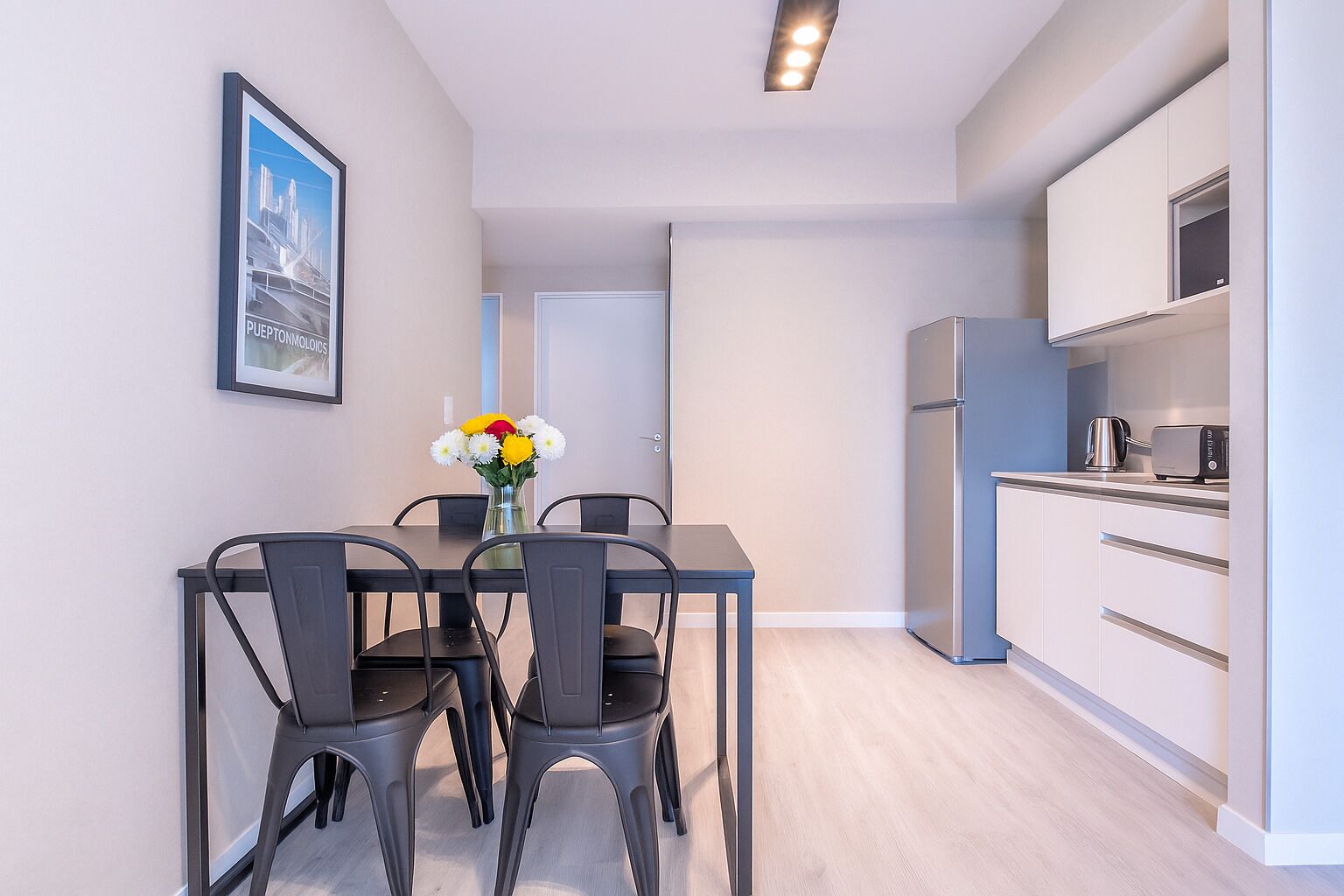 A modern kitchen with white cabinets and a stainless steel refrigerator. There is a dining table with four chairs and a vase with flowers.