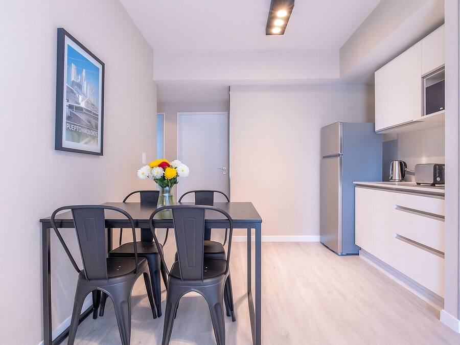 A modern kitchen with white cabinets and a stainless steel refrigerator. There is a dining table with four chairs and a vase with flowers.