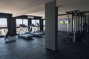 Een sportschool met loopbanden, een krachttrainingrek en een televisie waarop een sportevenement wordt vertoond.