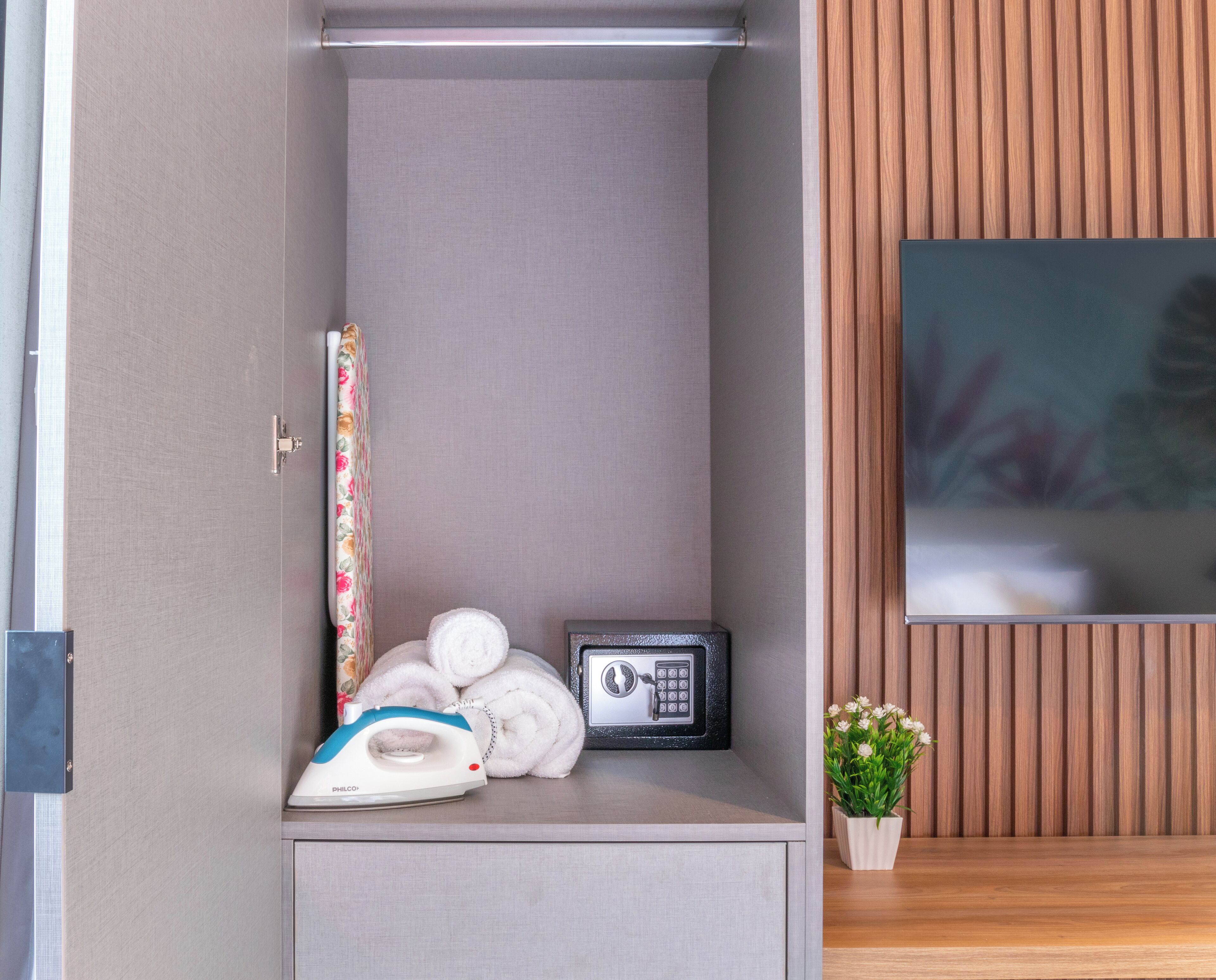 Una habitación pequeña con un armario gris, una plancha y tabla de planchar, una caja fuerte negra y una planta en maceta.