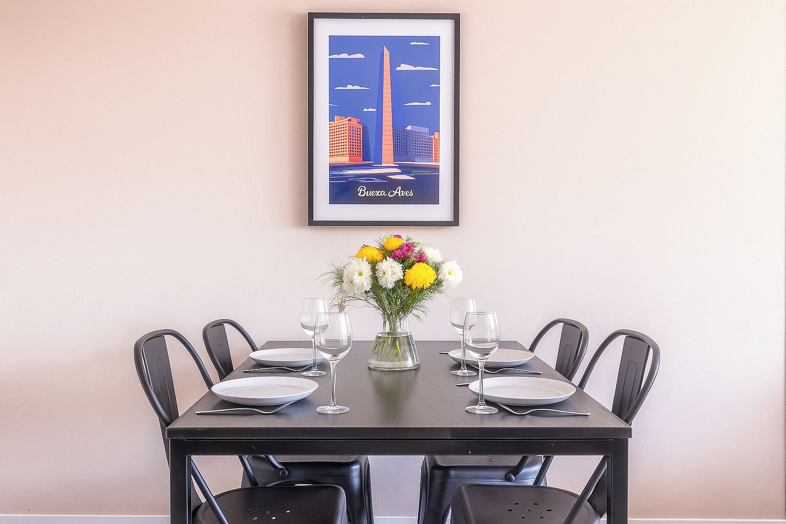 A dining table set for four with a framed cityscape poster, a vase of flowers, and four wine glasses.