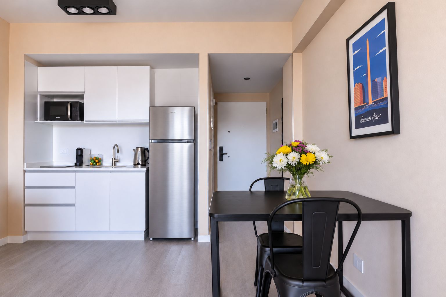 A compact kitchen with white cabinets, a stainless steel refrigerator, and a dining area with a black table and chairs.