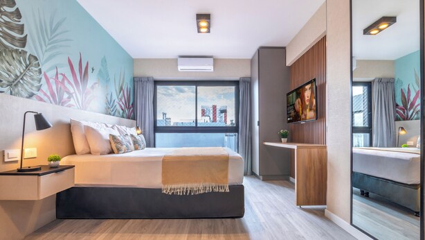 Una habitación de hotel moderna con una cama grande, una mesita de noche con una lámpara, una planta pequeña, un televisor y un ventanal con cortinas.