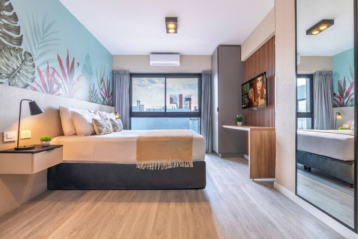 A modern hotel room with a large bed, a bedside table with a lamp, a small plant, a TV, and a large window with curtains.