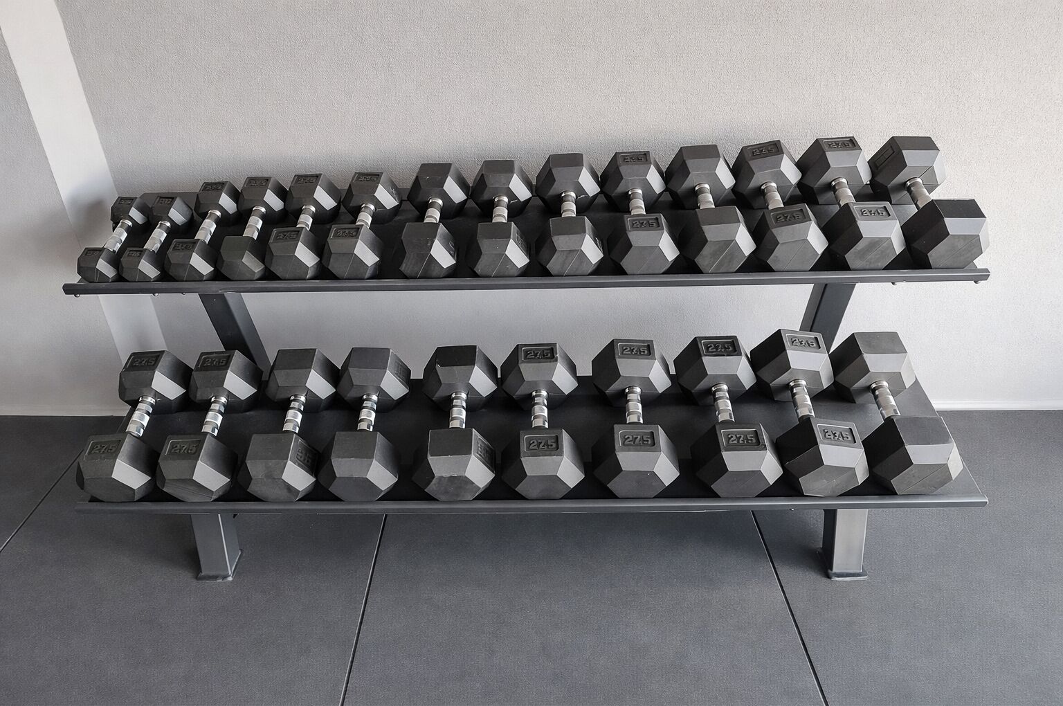 A rack of hexagonal dumbbells with weight markings, arranged on a metal stand against a plain wall.