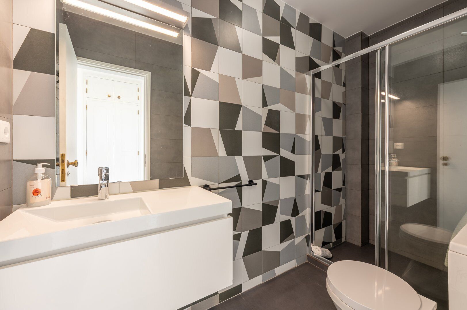 Une salle de bain moderne avec un mur recouvert de carreaux aux motifs géométriques, un lavabo blanc et une cabine de douche en verre.