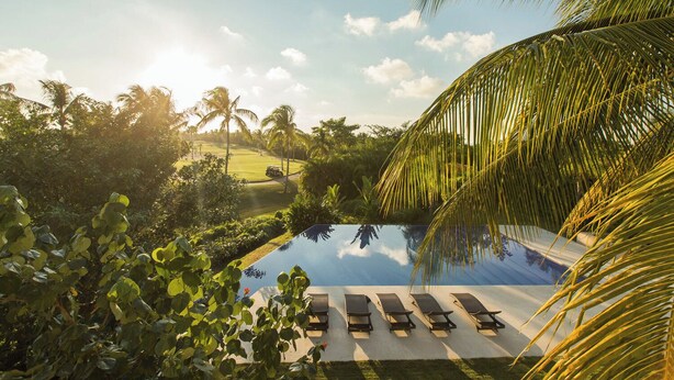 Une piscine bordée de chaises longues, entourée d’une végétation luxuriante et de palmiers, avec un terrain de golf en arrière-plan.