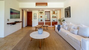 Un salon moderne avec un canapé blanc, une table basse et un coin repas comprenant une table et des chaises en bois. On y trouve également des étagères ornées d’objets décoratifs et une porte qui donne sur une autre pièce.