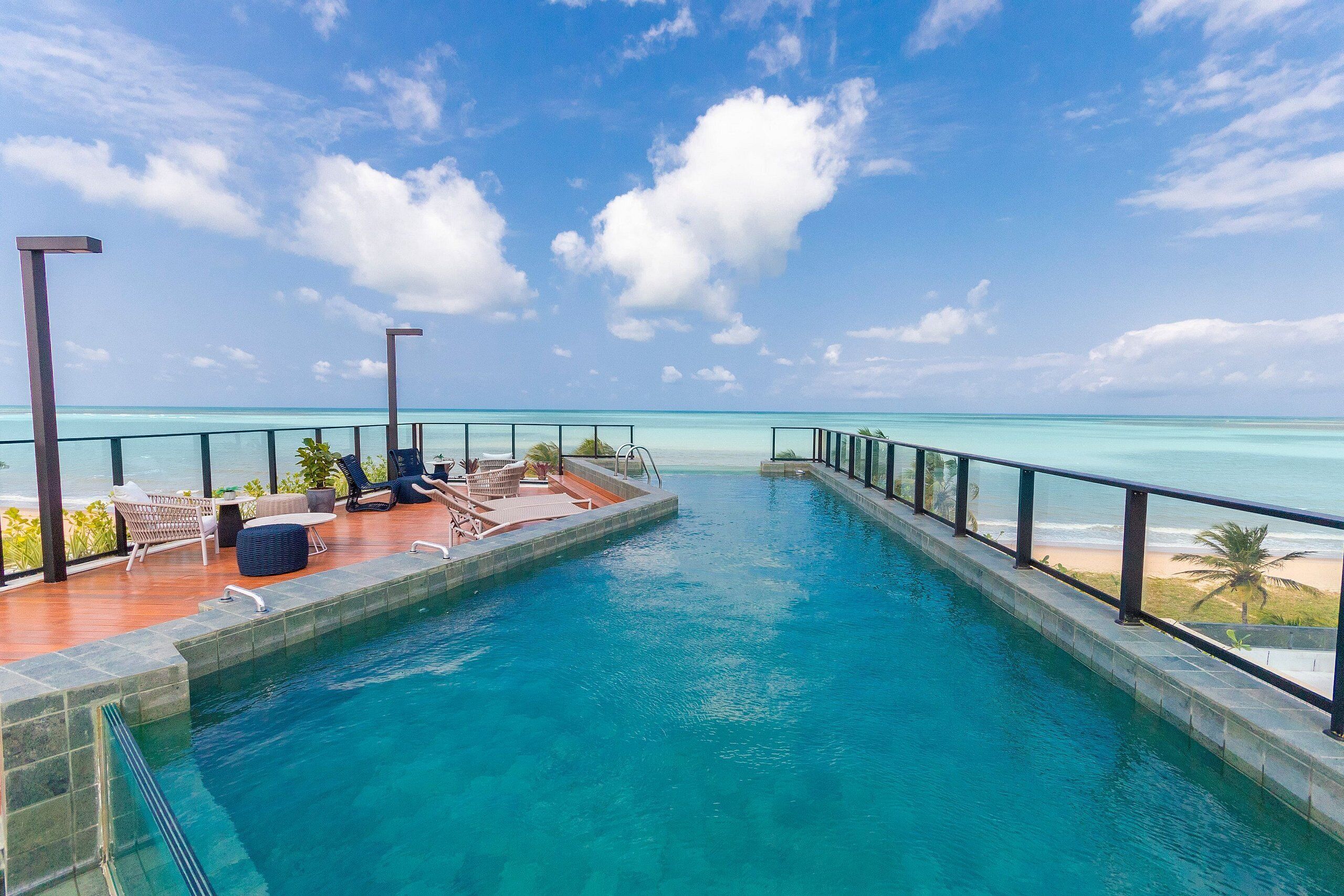 A pool with a view of the beach, lounge chairs, and a clear sky.
