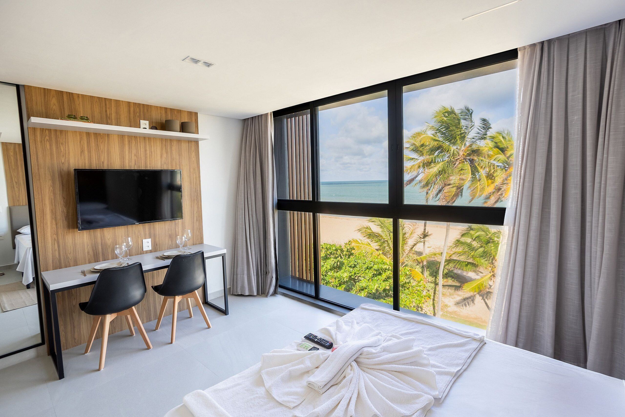 Habitación de hotel moderna con un ventanal grande, televisión de pantalla plana, zona de comedor y cama con vistas a palmeras.