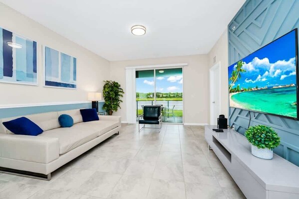 Ein modernes Wohnzimmer mit einem großen Flachbildfernseher, einem blauen Sofa mit Kissen, einer Topfpflanze und Blick auf einen Strand durch eine Schiebetür.