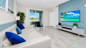 Una sala de estar moderna con un sofá color crema, cojines azules, un televisor de pantalla plana y vistas a una playa tropical a través de amplios ventanales.
