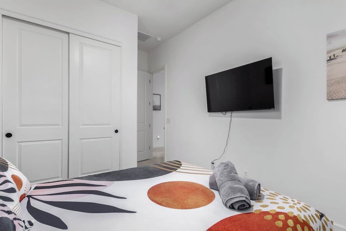 A modern hotel room with a large bed, a flat-screen TV mounted on the wall, and a hallway leading to a door.
