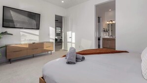A modern bedroom with a large bed, a wooden dresser, a flat-screen TV, and a bathroom visible through an open door. - Luxe 4BR Retreat w/ Pool, Hot Tub & Sunset Views (Goodyear)