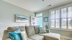 Een woonkamer met een hoekbank, een schilderij met een strandthema en een raam met jaloezieën.