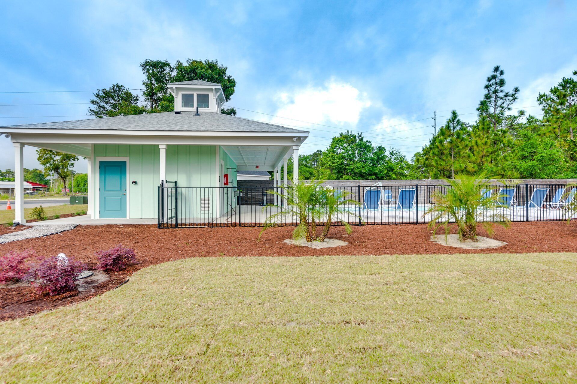 A single-story building with a covered porch, a small pool area with lounge chairs, and a fenced-in area with palm trees.