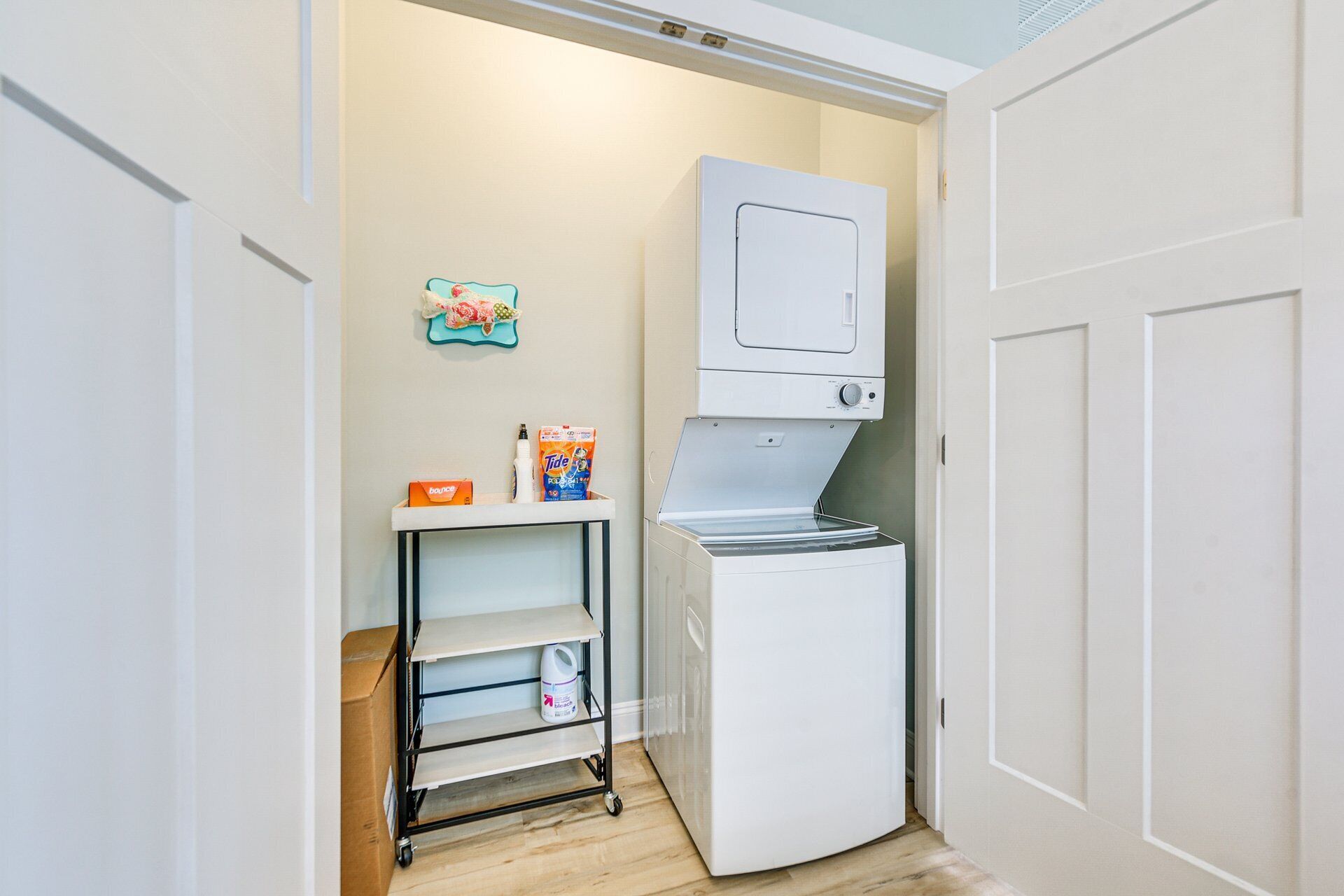 Un lavadero pequeño con lavadora y secadora blancas, un estante con productos de limpieza y una puerta que da a otra habitación.