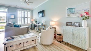 Een woonkamer met een bank, fauteuil en salontafel met tijdschriften. Een televisie hangt aan de muur, er is een plafondventilator en een raam met zonwering.