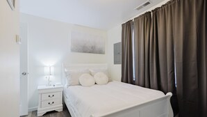 Ein Schlafzimmer mit einem weißen Bett, einem Nachttisch mit Lampe und einem Fenster mit Vorhängen.