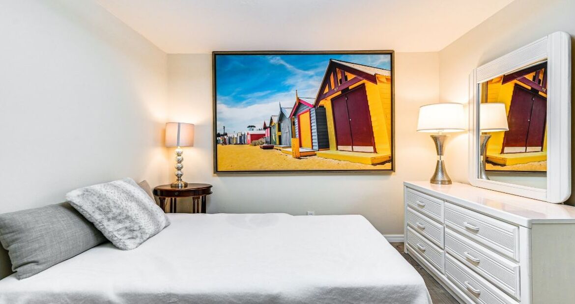 Ein Schlafzimmer mit Bett, Schminktisch, Nachttisch, Lampe und einem Bild bunter Strandhäuser.