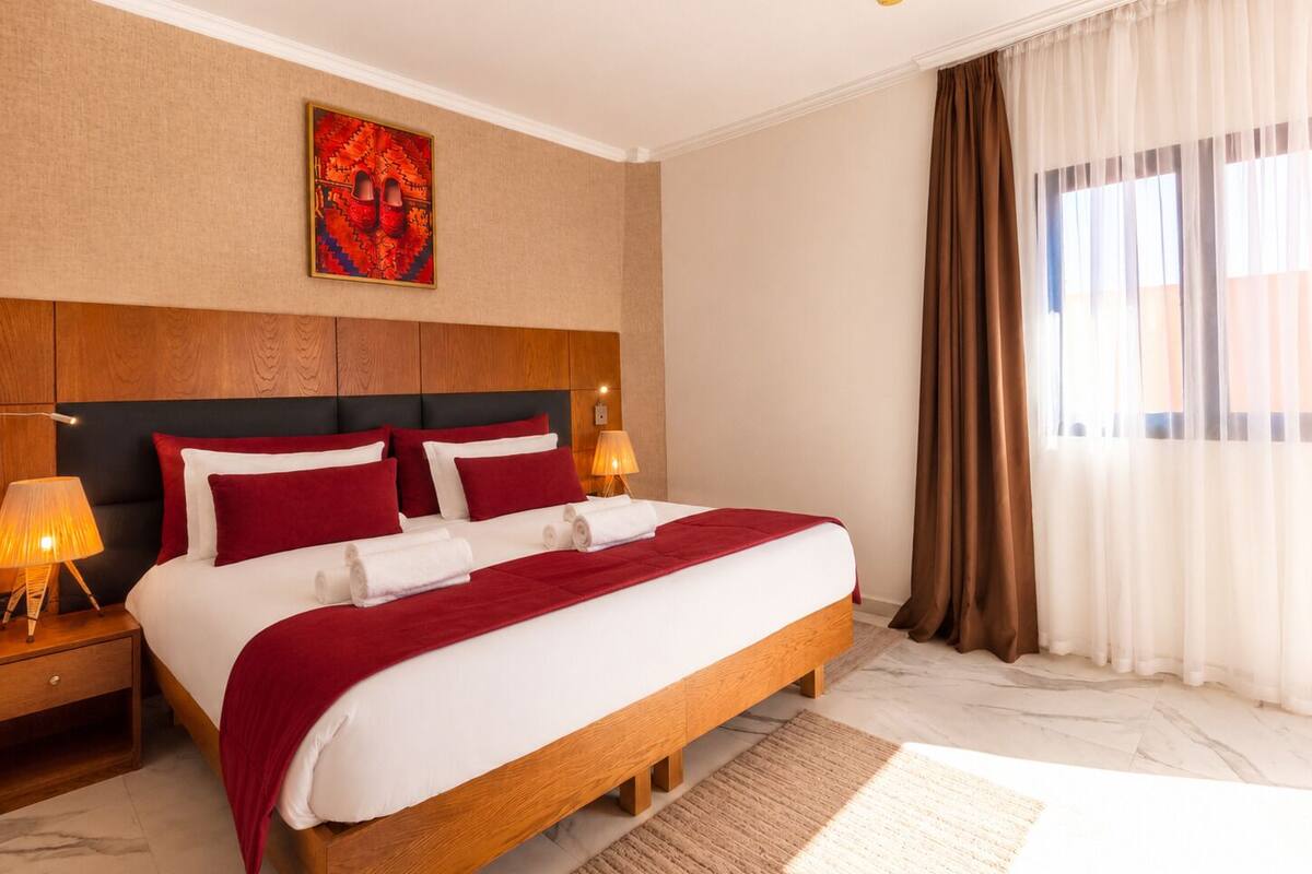 A hotel room with a large bed, wooden headboard, two bedside lamps, a red and white bedspread, a red throw blanket, a marble floor, a large window with curtains, and a wall art piece.