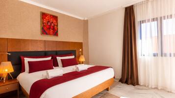 Habitación de hotel con una cama grande, cabecero de madera, dos lámparas de noche, colcha blanca y roja, manta roja, piso de mármol, un ventanal con cortinas y una obra de arte en la pared.