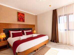 A hotel room with a large bed, wooden headboard, two bedside lamps, a red and white bedspread, a red throw blanket, a marble floor, a large window with curtains, and a wall art piece. - Ourika Camp Suites Hotel & Resort (Marrakech)