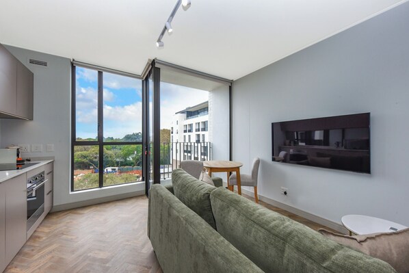 A modern apartment with a large sliding glass door leading to a balcony, a green sofa, a small dining table, and a TV mounted on the wall.