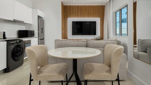 Une cuisine moderne avec des meubles blancs, un four encastré et un lave-linge. On y trouve une table de salle à manger ronde, accompagnée de quatre chaises, et un grand téléviseur à écran plat fixé à un mur en bois.