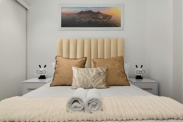 Una cama bien hecha con cabecera, dos mesitas de noche con almohadones decorativos y un cuadro en la pared.