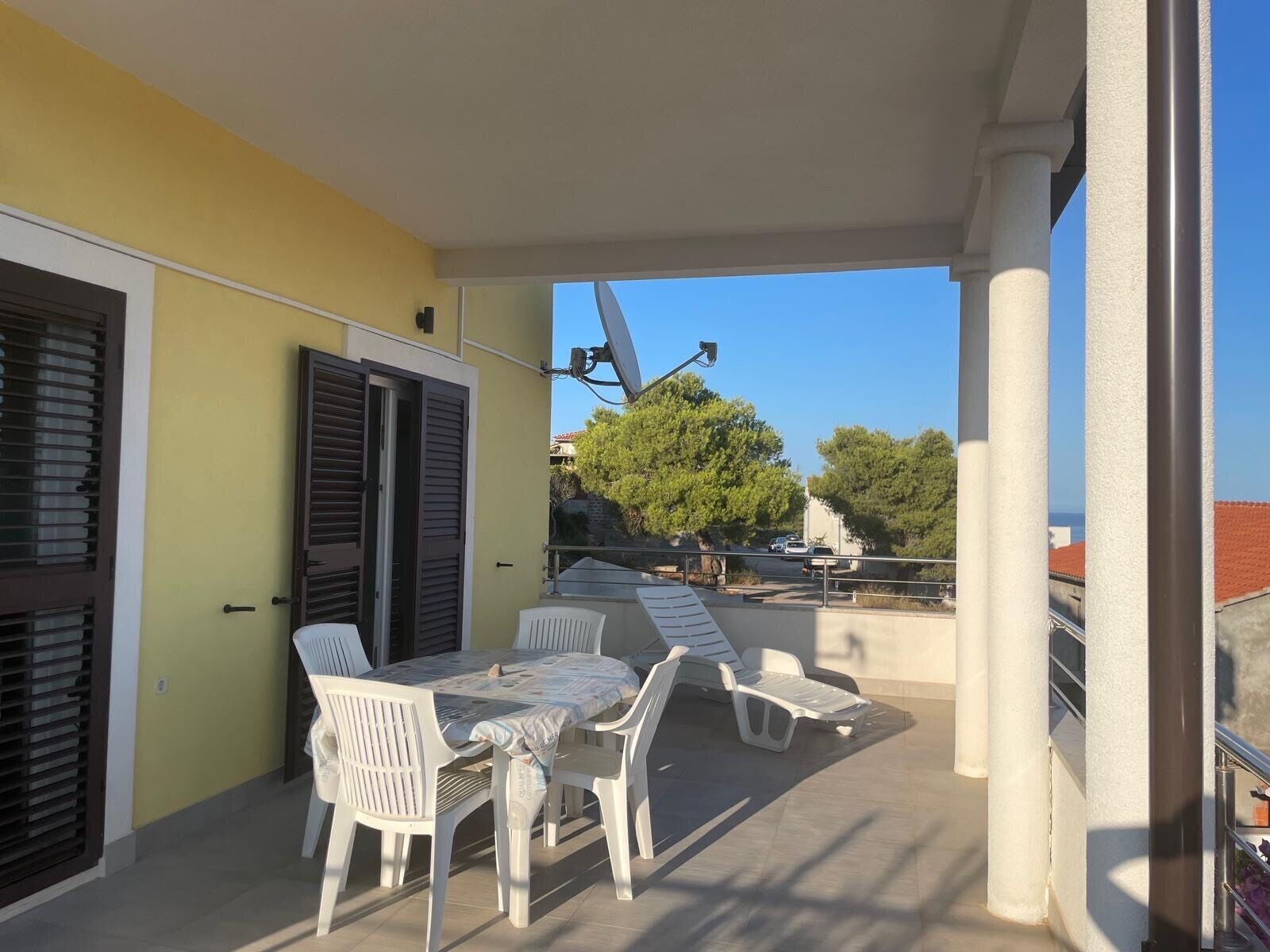 Un patio exterior con una mesa y sillas, una puerta con contraventanas y vista a la calle y árboles.