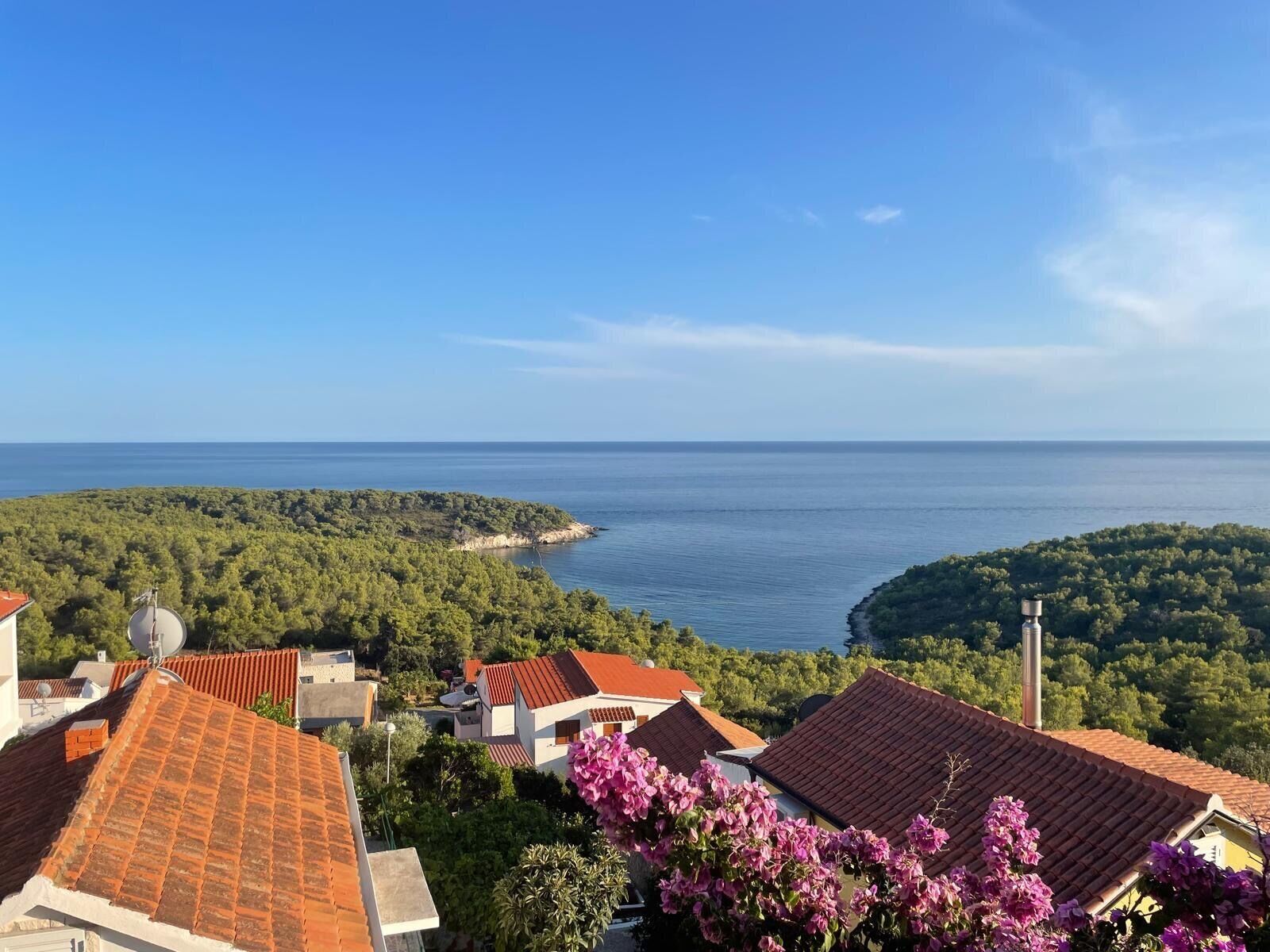 A coastal village with terracotta-roofed houses, lush greenery, and a view of the sea.