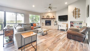 A spacious living room with a stone fireplace, a ceiling fan, and a large window overlooking a deck.
