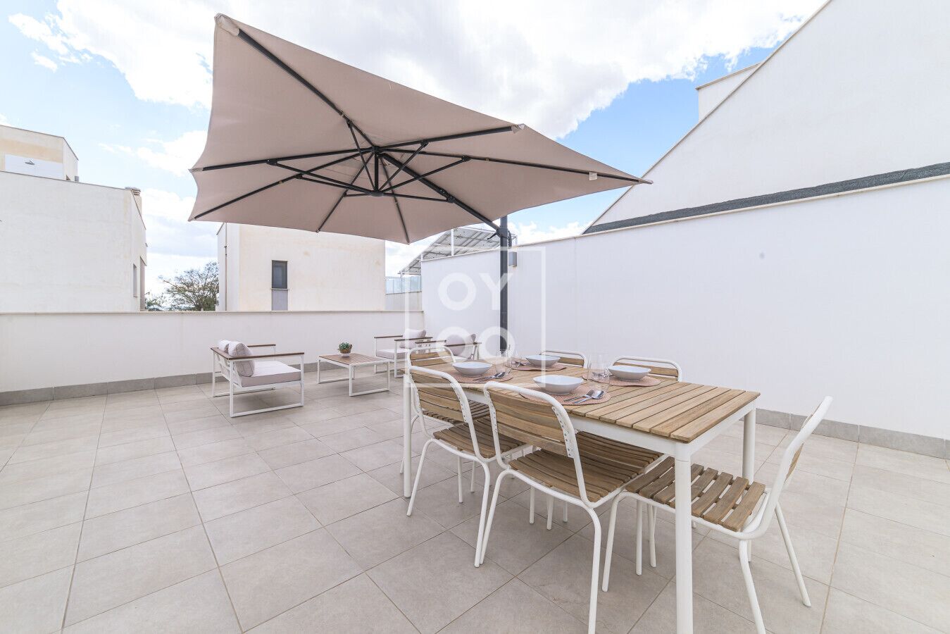 A rooftop patio with a wooden table and chairs, a large umbrella, and a white wall in the background.