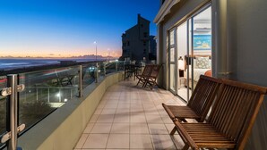 Ein Balkon mit Meerblick, im Hintergrund ein Gebäude und ein Tisch mit Stühlen.