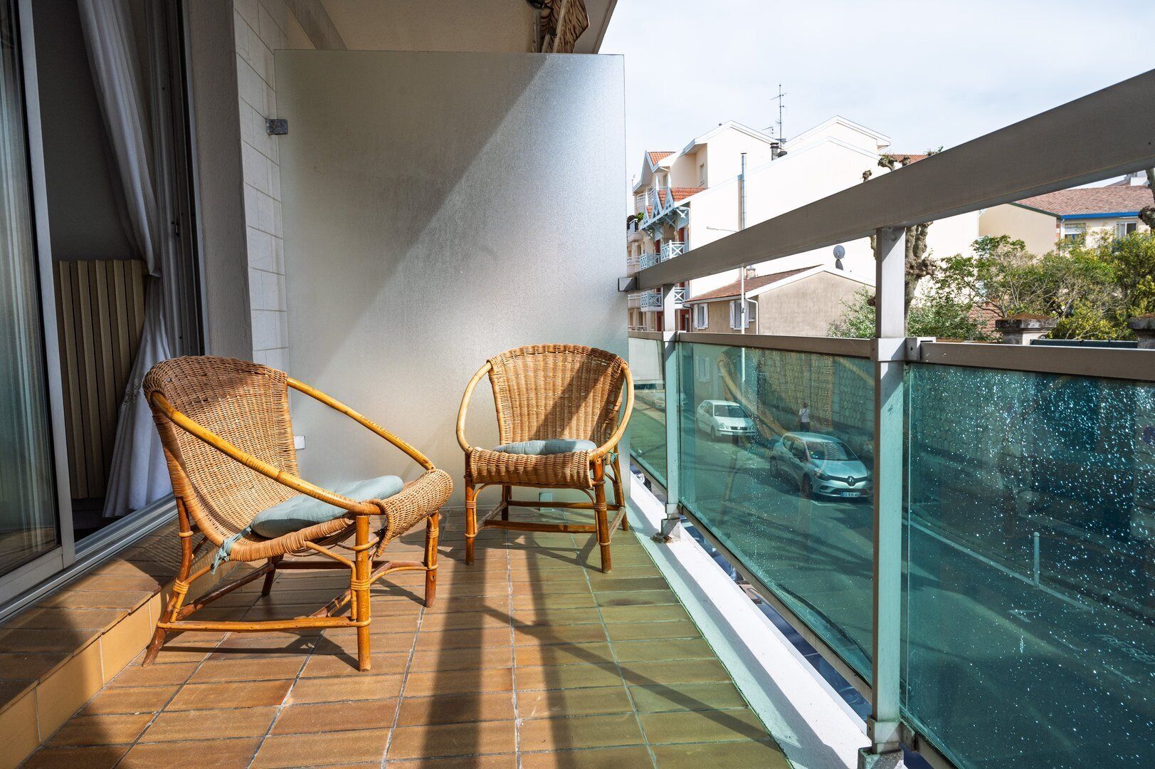 A balcony with two wicker chairs, a glass railing, and a view of a street with cars.