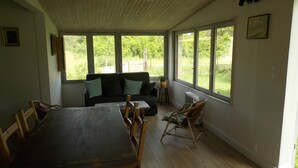 A room with a wooden dining table, a sofa, and a rocking chair. There are large windows providing a view of greenery outside.