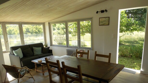 A dining area with a wooden table and chairs, a sofa with cushions, and large windows offering a view of the outdoors.