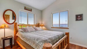 Quarto com estrutura de cama de madeira, duas janelas com persianas, um espelho redondo e uma pequena mesa de cabeceira.